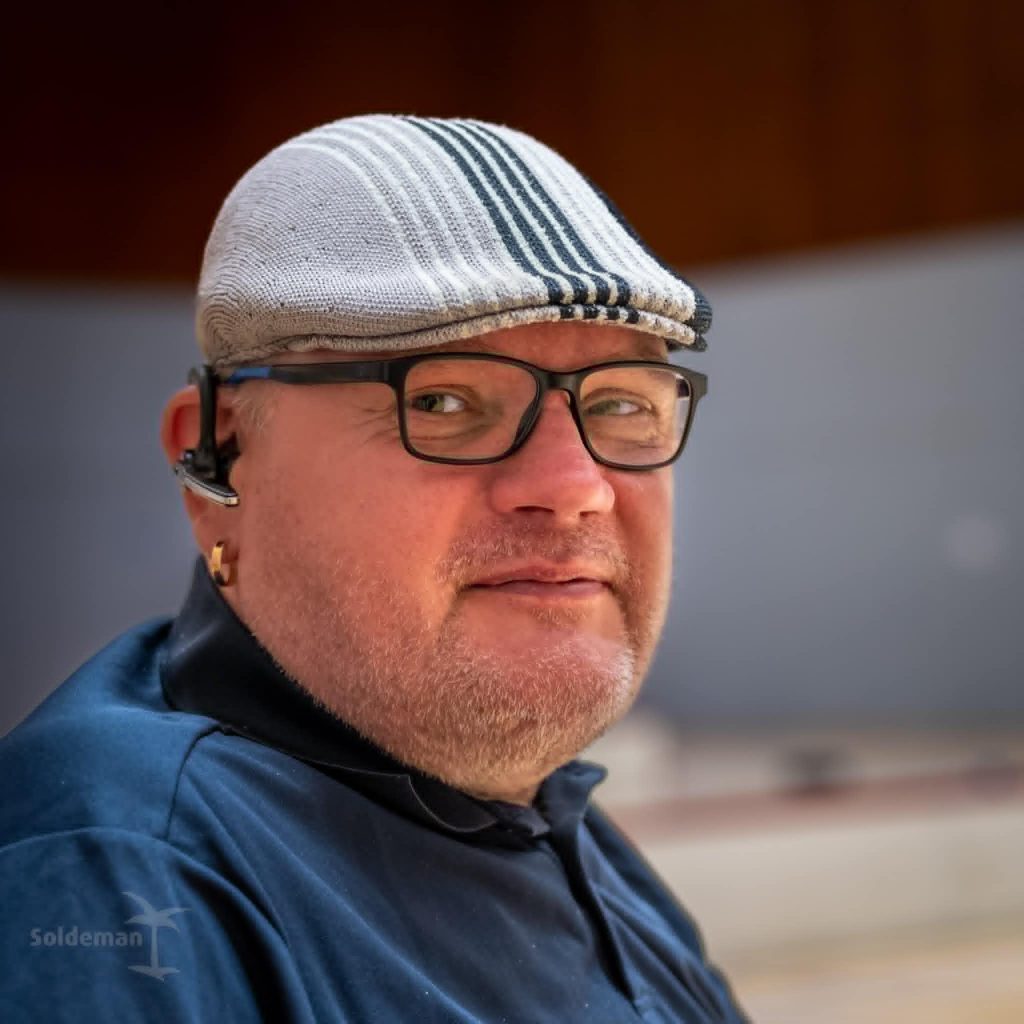 Silmälasipäinen mies harmaa flat cap -tyylinen lippalakki päässä.